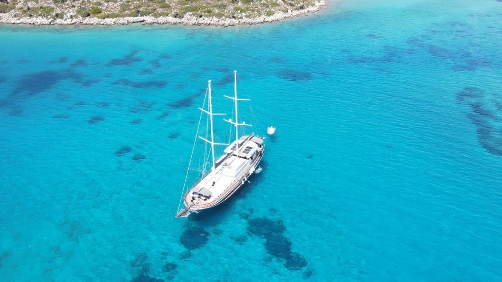 Aerial view of a sailboat anchored in clear turquoise water near a rocky coastline, with patches of underwater rocks visible through the water.