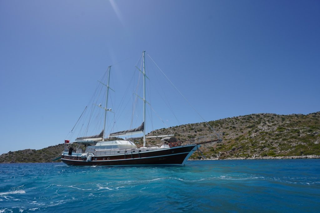 A large sailboat with two masts floats on clear blue water near a rocky, green coastline under a clear, bright blue sky.