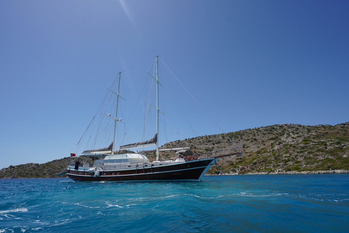 A large sailboat with two masts floats on clear blue water near a rocky, green coastline under a clear, bright blue sky.