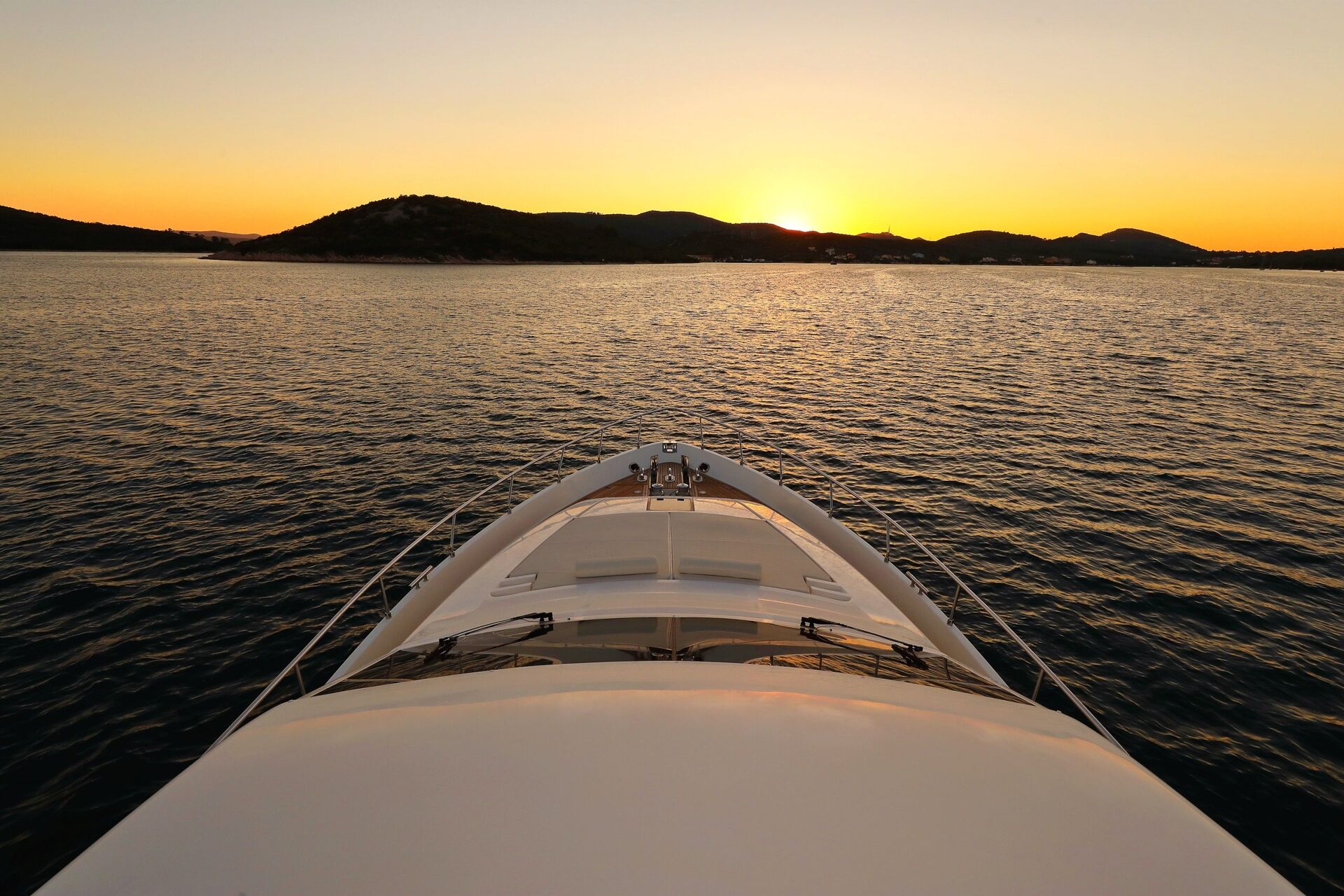 Un yate navega por aguas tranquilas al atardecer, con el sol poniéndose tras unas colinas lejanas y el cielo resplandeciente de naranja y amarillo. En primer plano se ve la proa del yate.