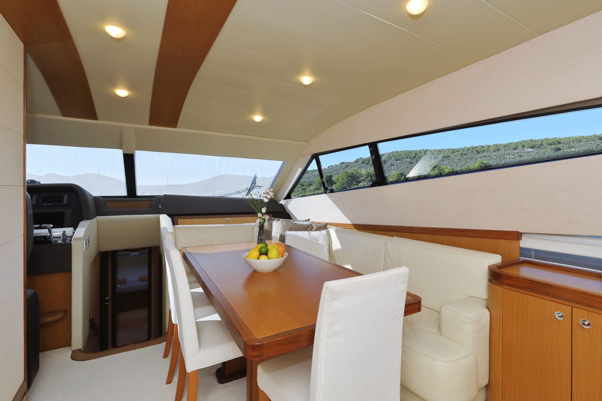 Interior de un yate moderno con una mesa de comedor de madera, sillas blancas tapizadas, un sofá blanco y un frutero; grandes ventanales ofrecen vistas al mar y a las verdes colinas del exterior.