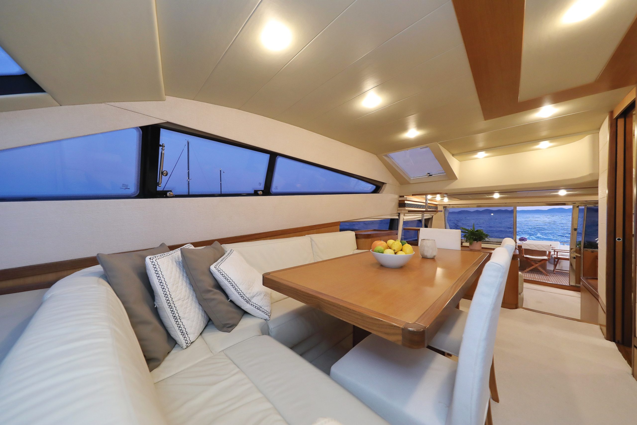 Moderno interior de yate con asientos de cuero color crema, mesa de madera coronada con un frutero y cojines decorativos; grandes ventanales muestran una vista del mar y los veleros al atardecer. Las cálidas luces del techo iluminan el espacio.