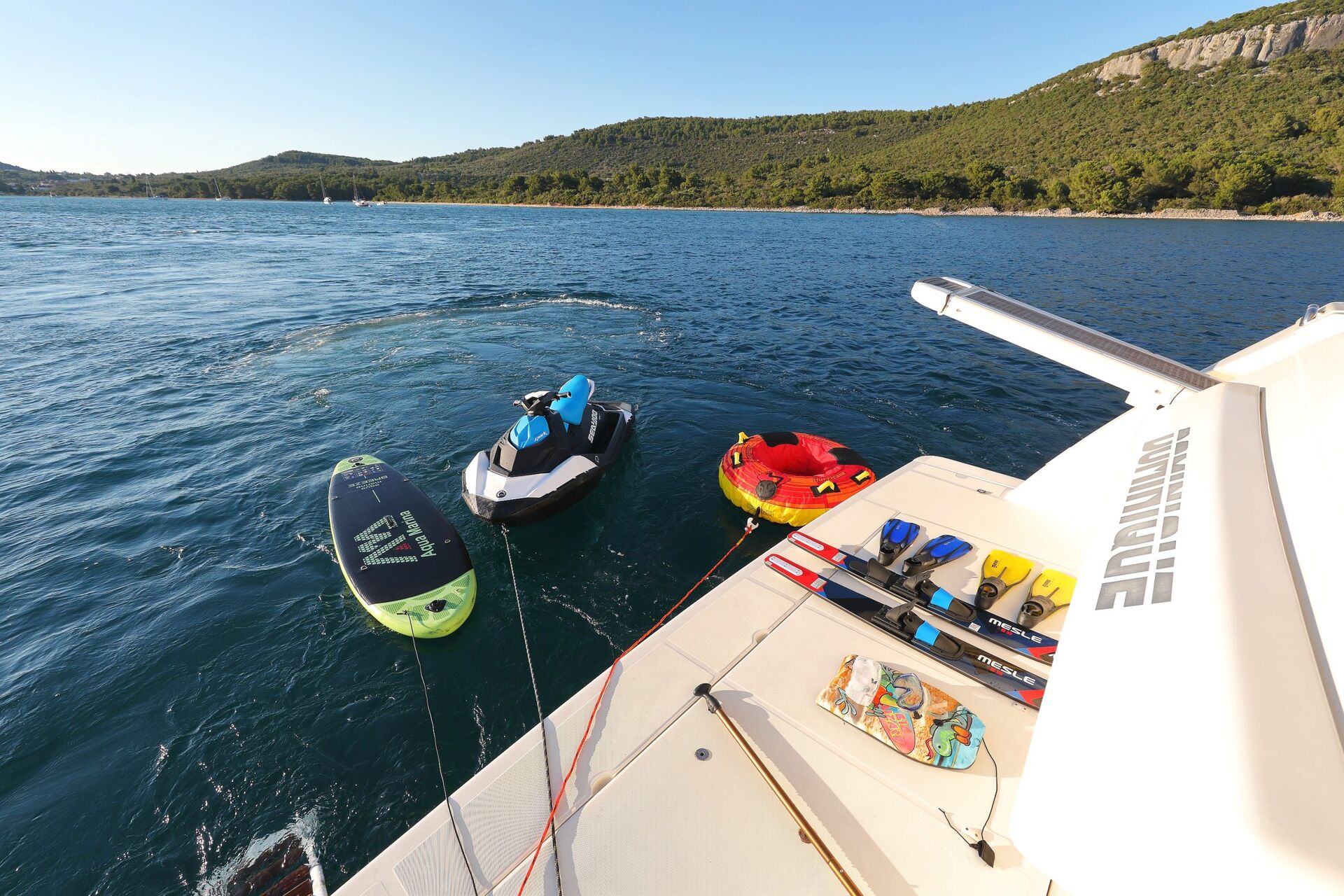 Una vista desde la cubierta de un barco muestra una tabla de paddle, una moto acuática y dos tubos inflables flotando en aguas azules cerca de una costa boscosa bajo un cielo despejado. En la cubierta hay varios equipos de deportes acuáticos.