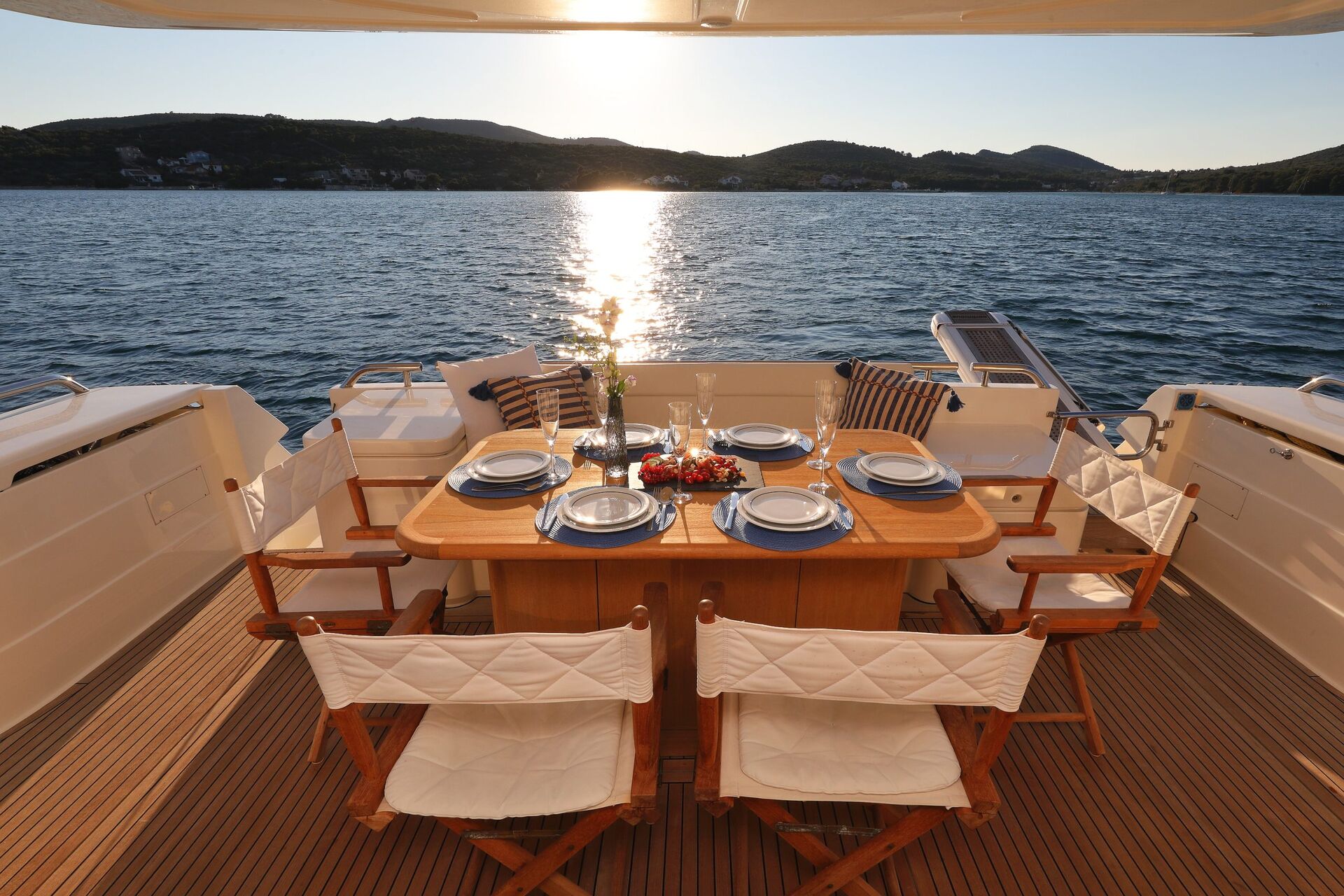 Una mesa de comedor para seis personas con sillas blancas está en la cubierta de un yate, con vistas a aguas tranquilas y colinas al atardecer, con la luz del sol reflejándose en el mar.