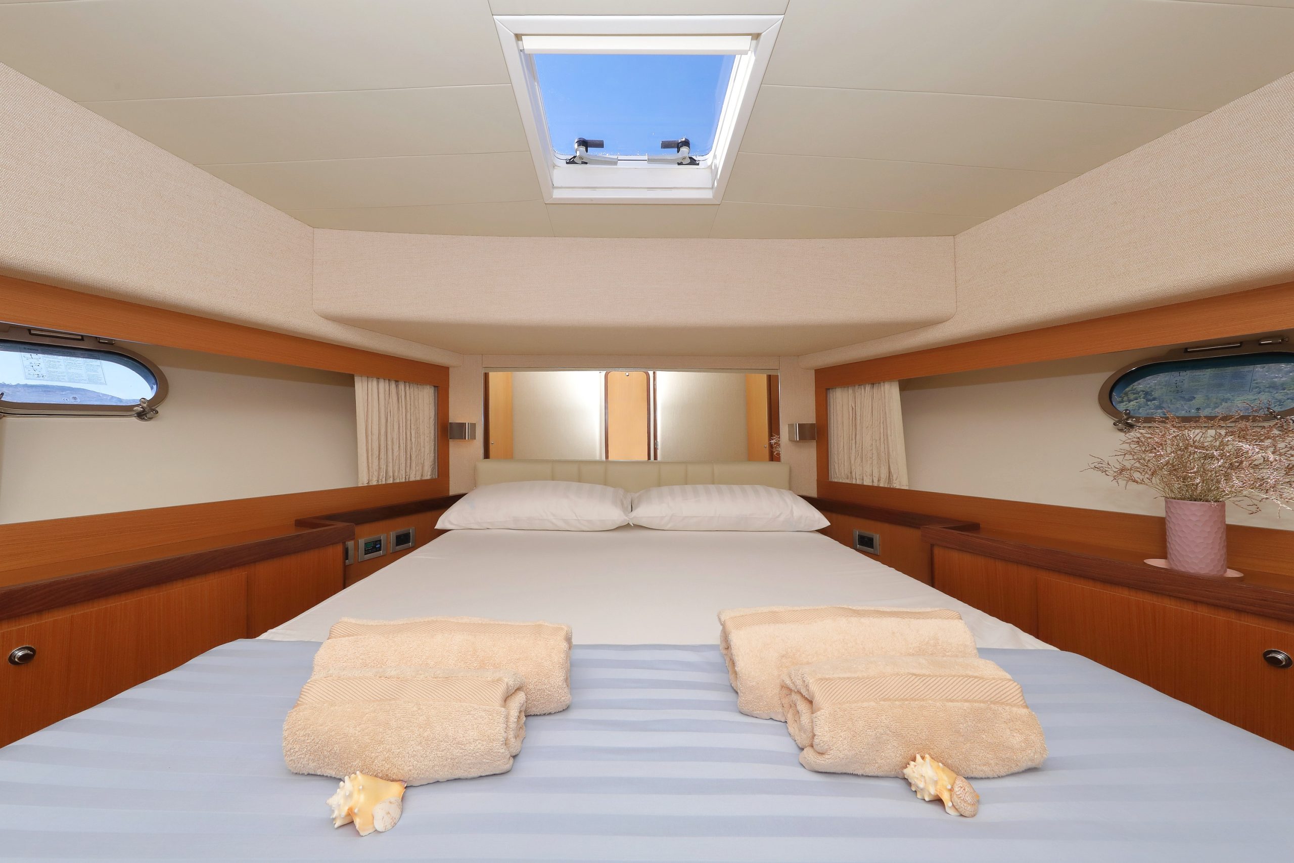 Una cama bien hecha en una acogedora cabaña con dos pilas de toallas dobladas, conchas marinas encima y una claraboya encima que deja entrar la luz natural. Hay pequeñas ventanas y una planta en un estante de madera.