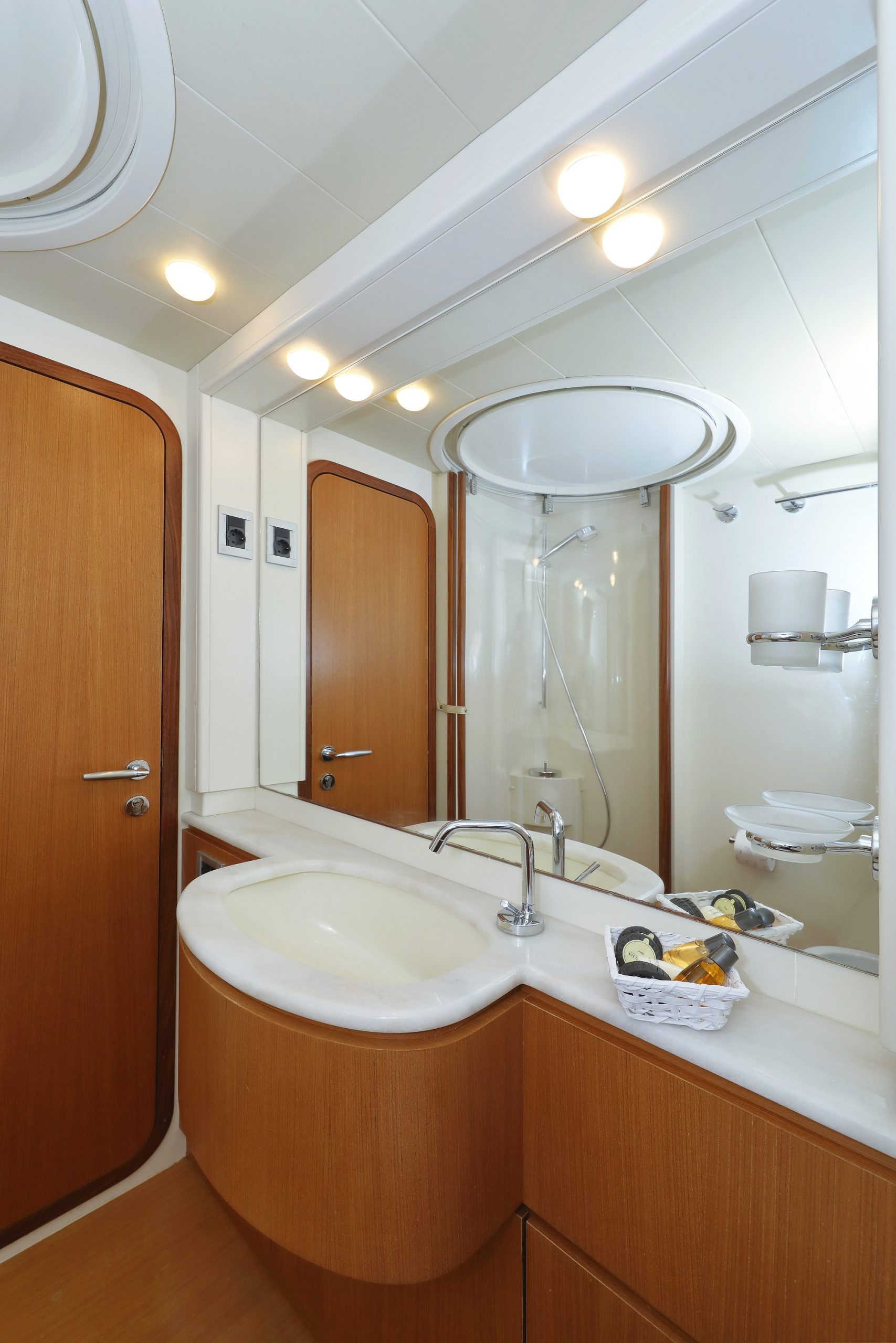 Un cuarto de baño moderno con puerta de madera, espejo grande, lavabo con encimera curva y cabina de ducha con puertas de cristal. Hay artículos de aseo en la encimera y luces brillantes en el techo.