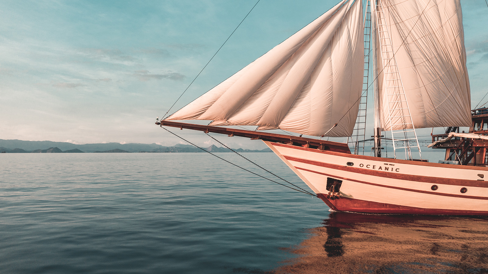 A wooden sailboat with white sails, named Oceanic, is anchored on calm blue water near distant mountains under a clear sky.