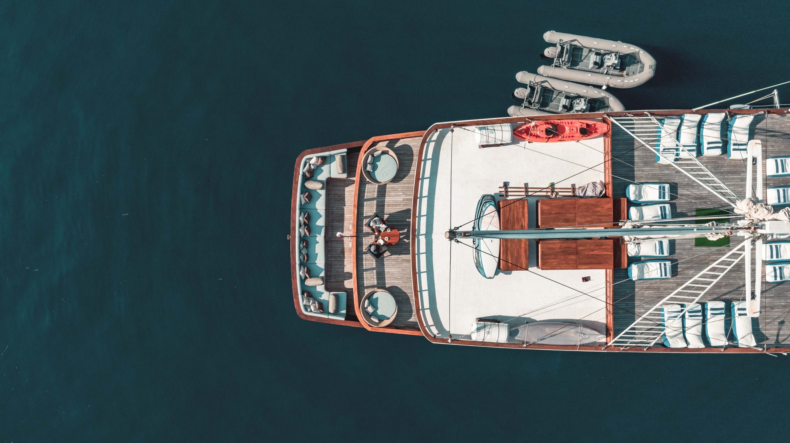 Aerial view of a yacht on calm dark blue water, featuring sun loungers, seating areas, a red kayak, and two small inflatable boats attached to the rear. Two people are relaxing on the upper deck.