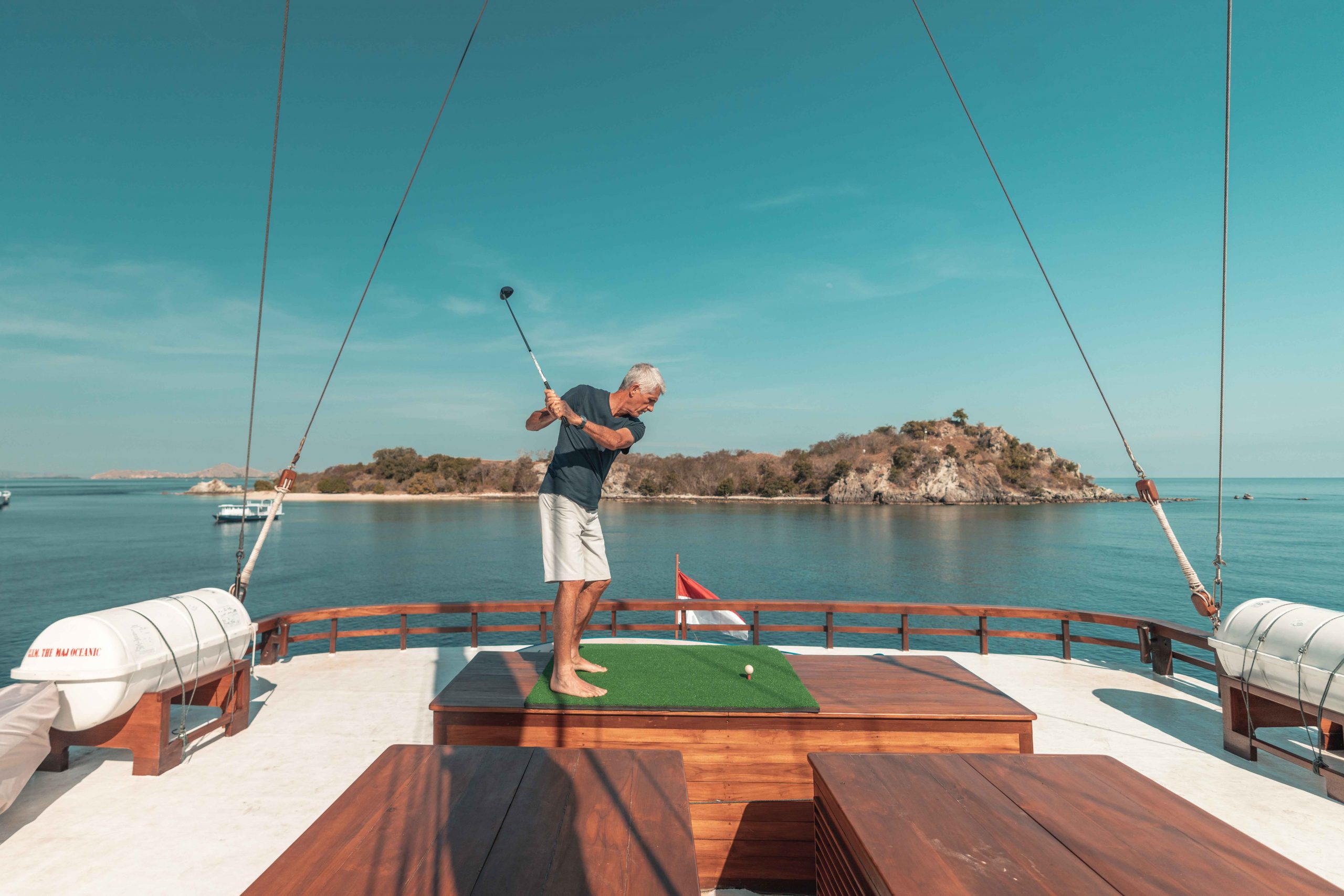A man swings a golf club on the deck of a boat, aiming at a golf ball on a small green mat, with calm blue water and a small island in the background.
