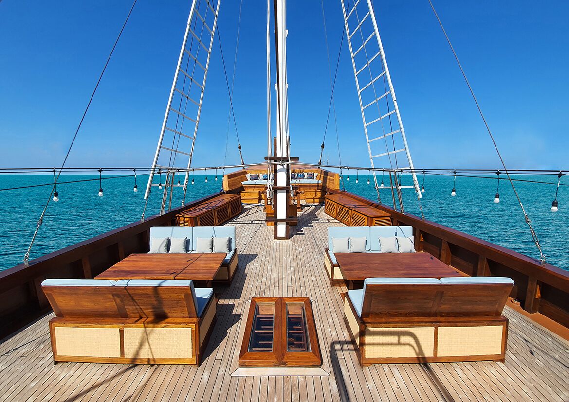 Spacious wooden deck of a yacht with cushioned seating areas, wooden tables, and clear railings overlooking calm, blue ocean waters under a clear sky.