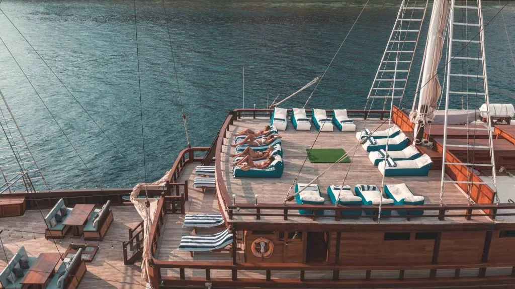 A group of people relax on cushioned lounge chairs on the deck of a wooden yacht, anchored in calm blue water, with sunlight illuminating the scene.