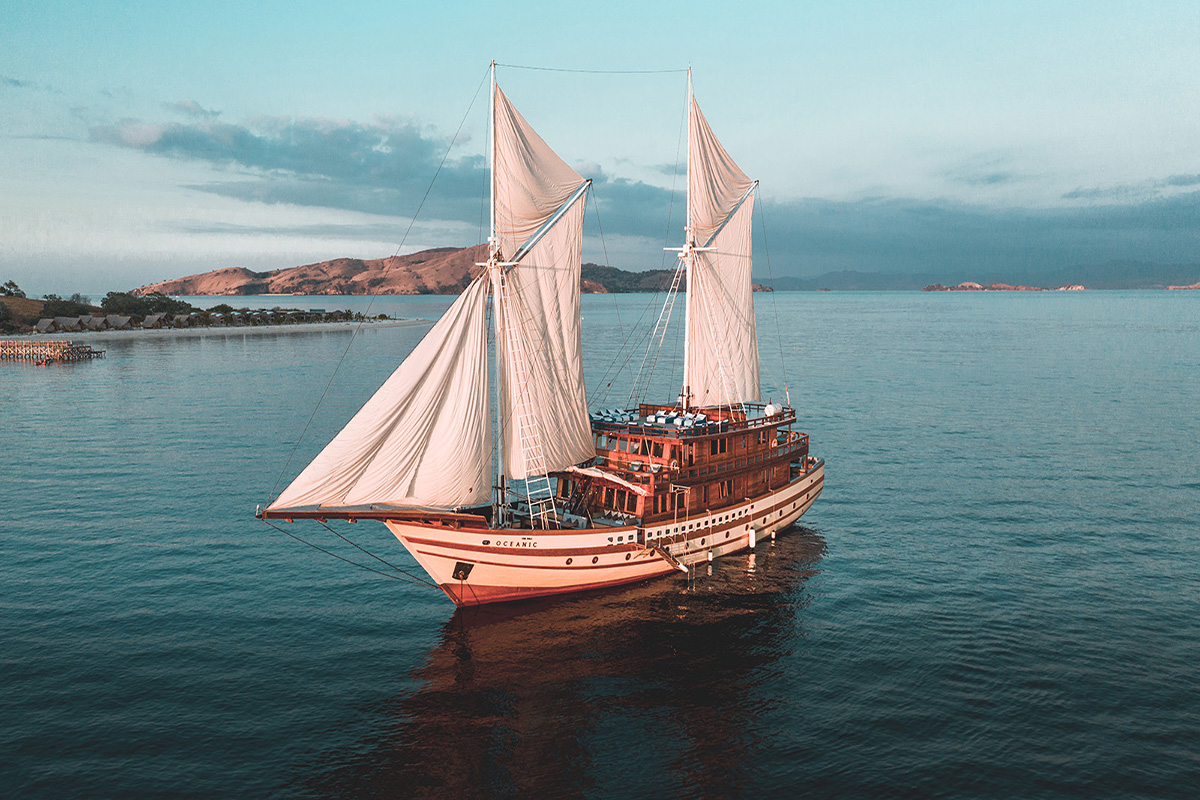 A traditional wooden sailing ship with white sails glides on calm blue water near a coastline with hills under a clear sky.