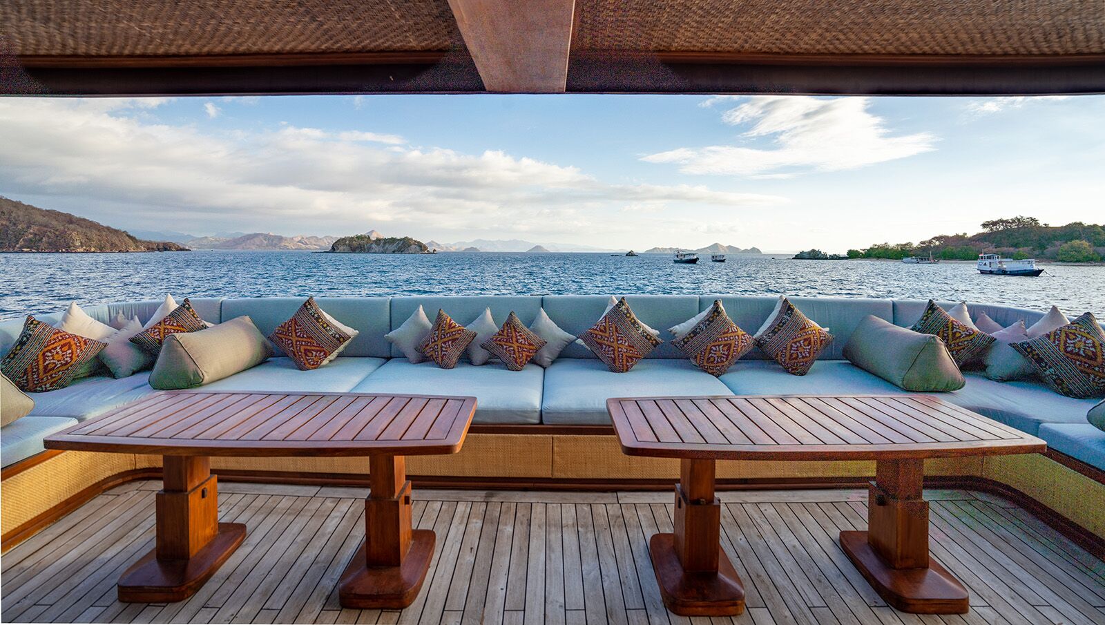 Spacious yacht deck with two wooden tables, cushioned bench seating, and patterned pillows, overlooking calm blue water and distant islands under a partly cloudy sky.