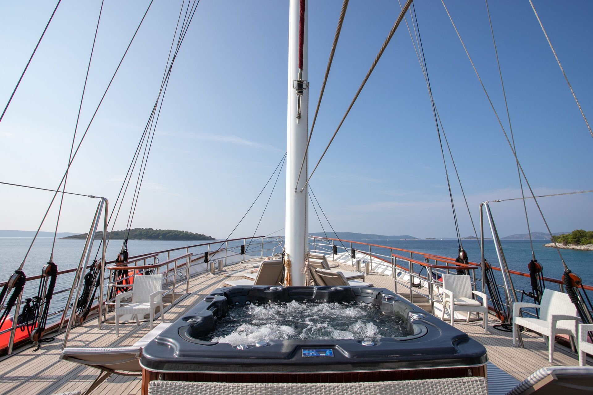 A luxury yacht deck featuring a bubbling hot tub, white chairs, and wooden railings awaits you—perfect for a serene yacht charter with views of calm blue sea and distant green islands under a clear sky.