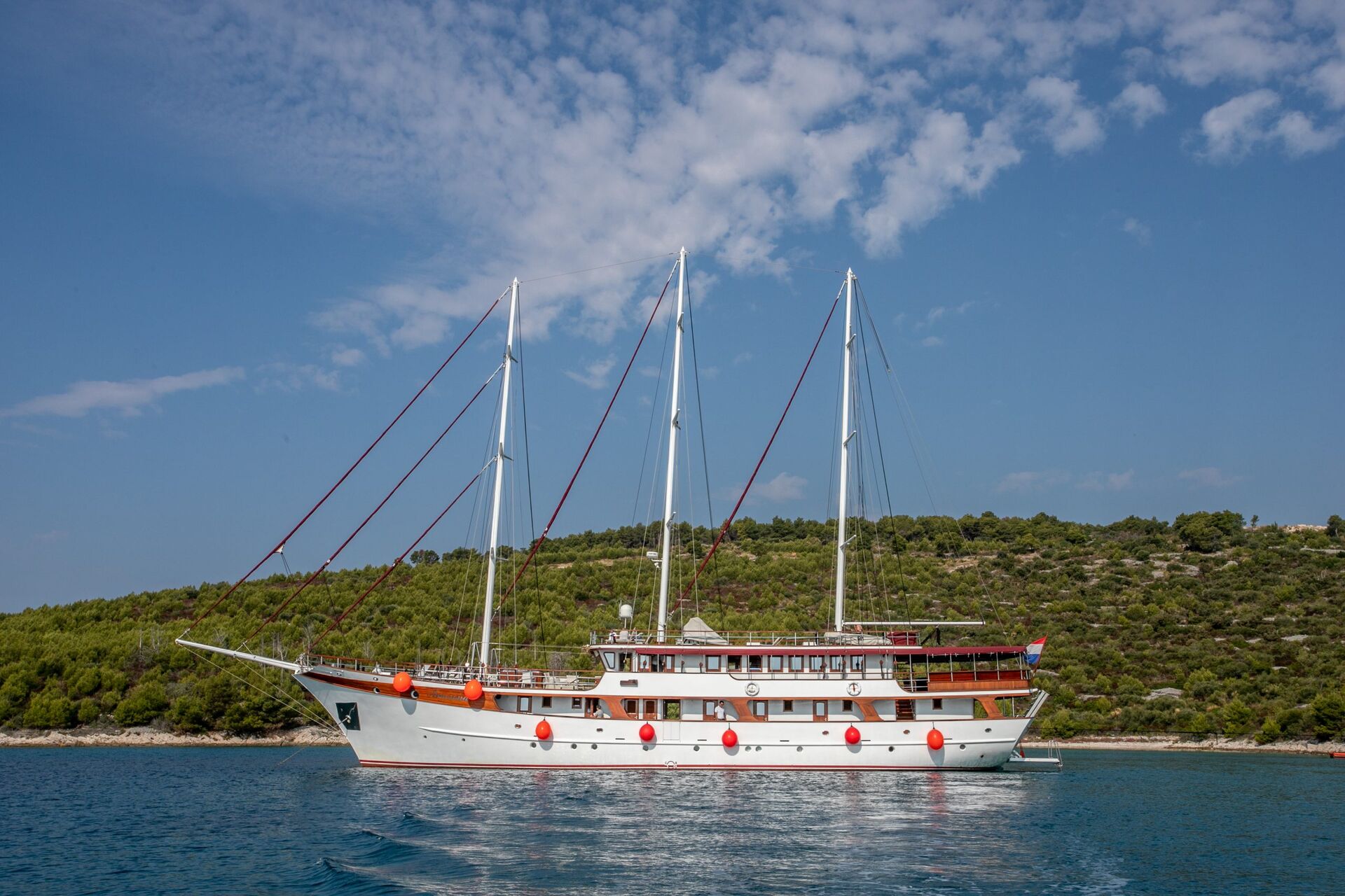 A large white three-masted sailing ship with red fenders floats on calm blue water, perfect for yacht rental, with a green, tree-covered shoreline and a partly cloudy sky in the background.