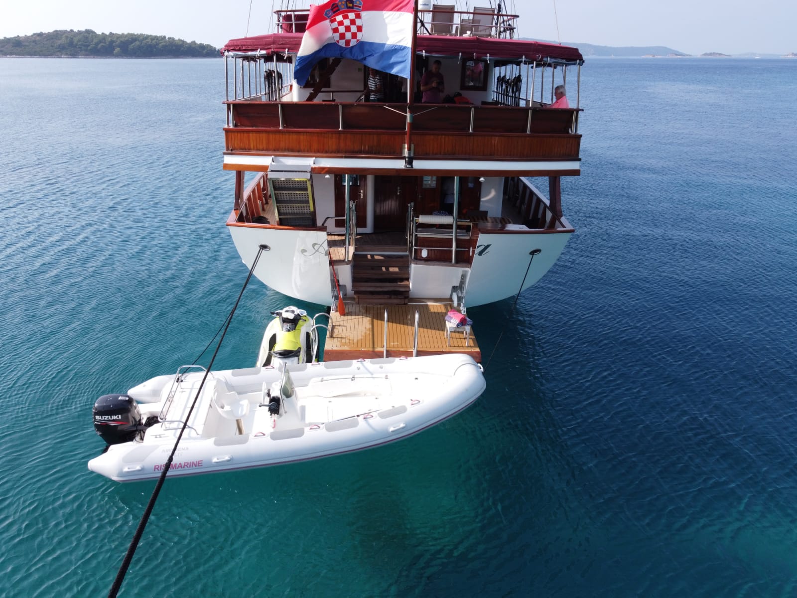 A white inflatable boat is tied to the stern of a wooden yacht with a Croatian flag, floating on calm blue water—an inviting scene for those considering yachts for charter, with distant land visible in the background.