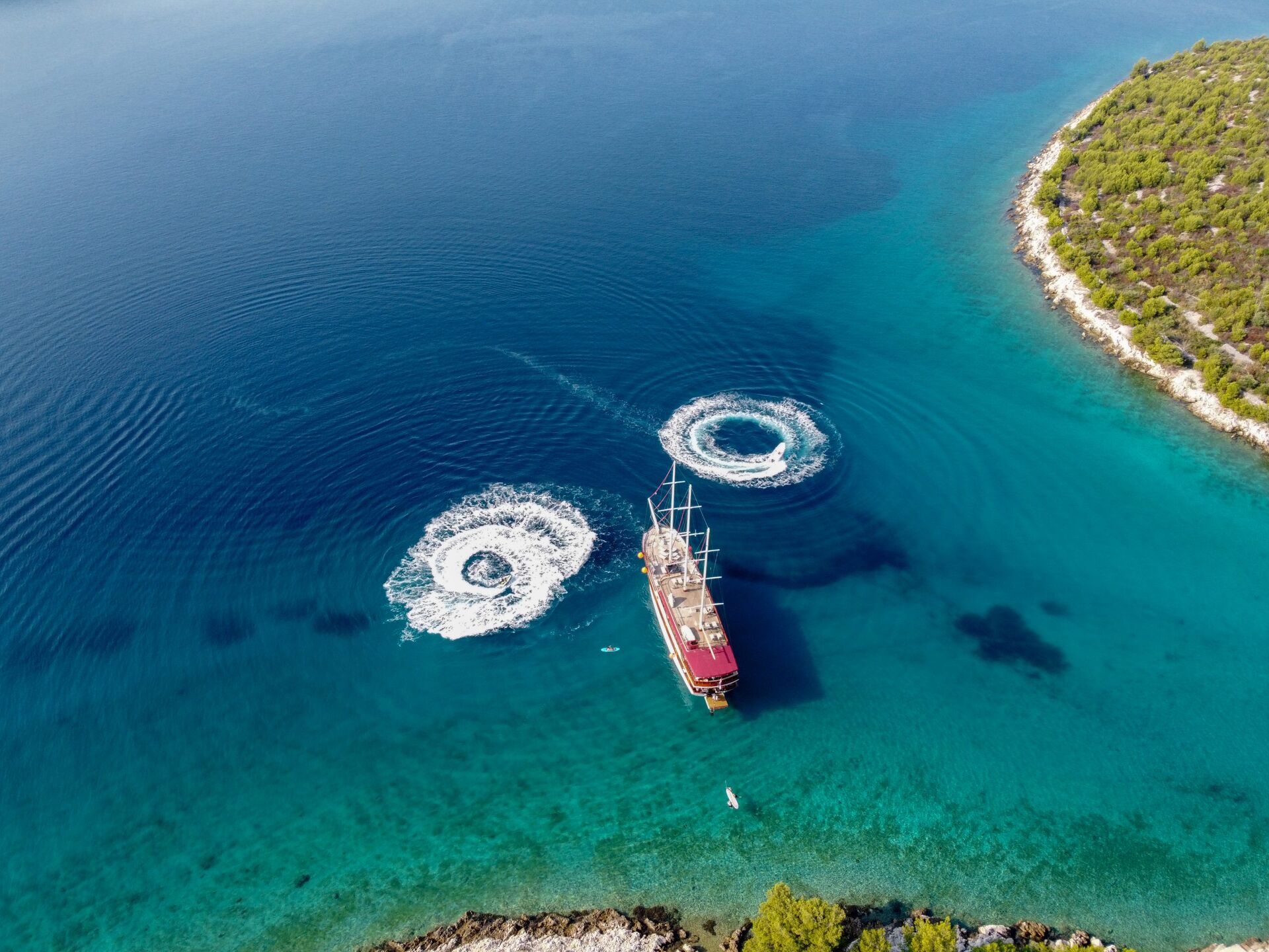 Aerial view of a wooden sailing ship in clear blue water near a rocky, tree-covered coastline, with two circular patterns from boats nearby—perfect for those seeking unique yacht charter experiences.