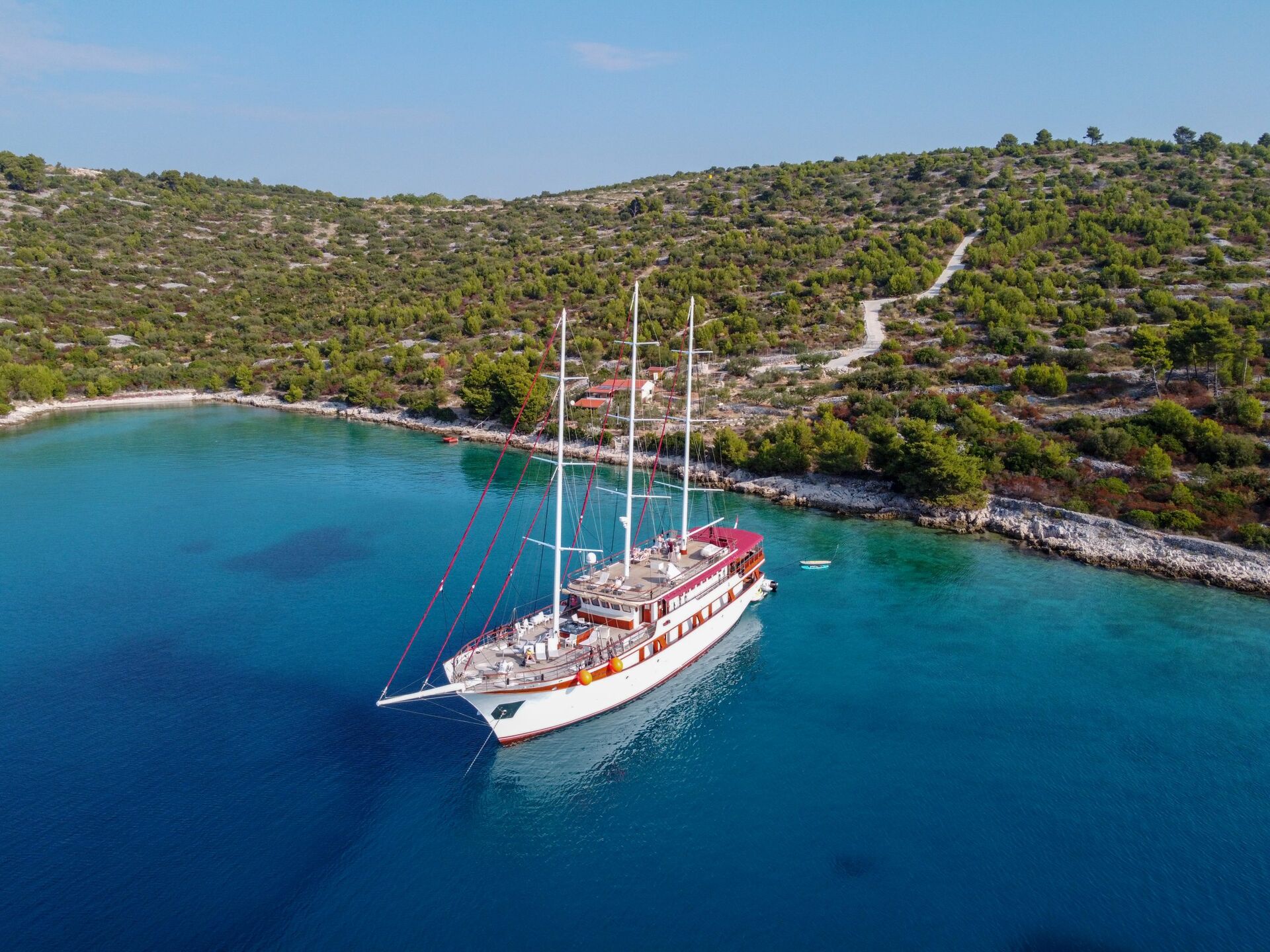 A large white sailing yacht, available for yacht charter, is anchored in a calm blue bay near a hilly green coastline with scattered trees and a winding path under a clear sky.