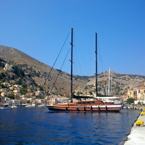 A large wooden sailboat with two masts is anchored in a calm harbor, with hillside houses and mountains in the background under a clear blue sky.