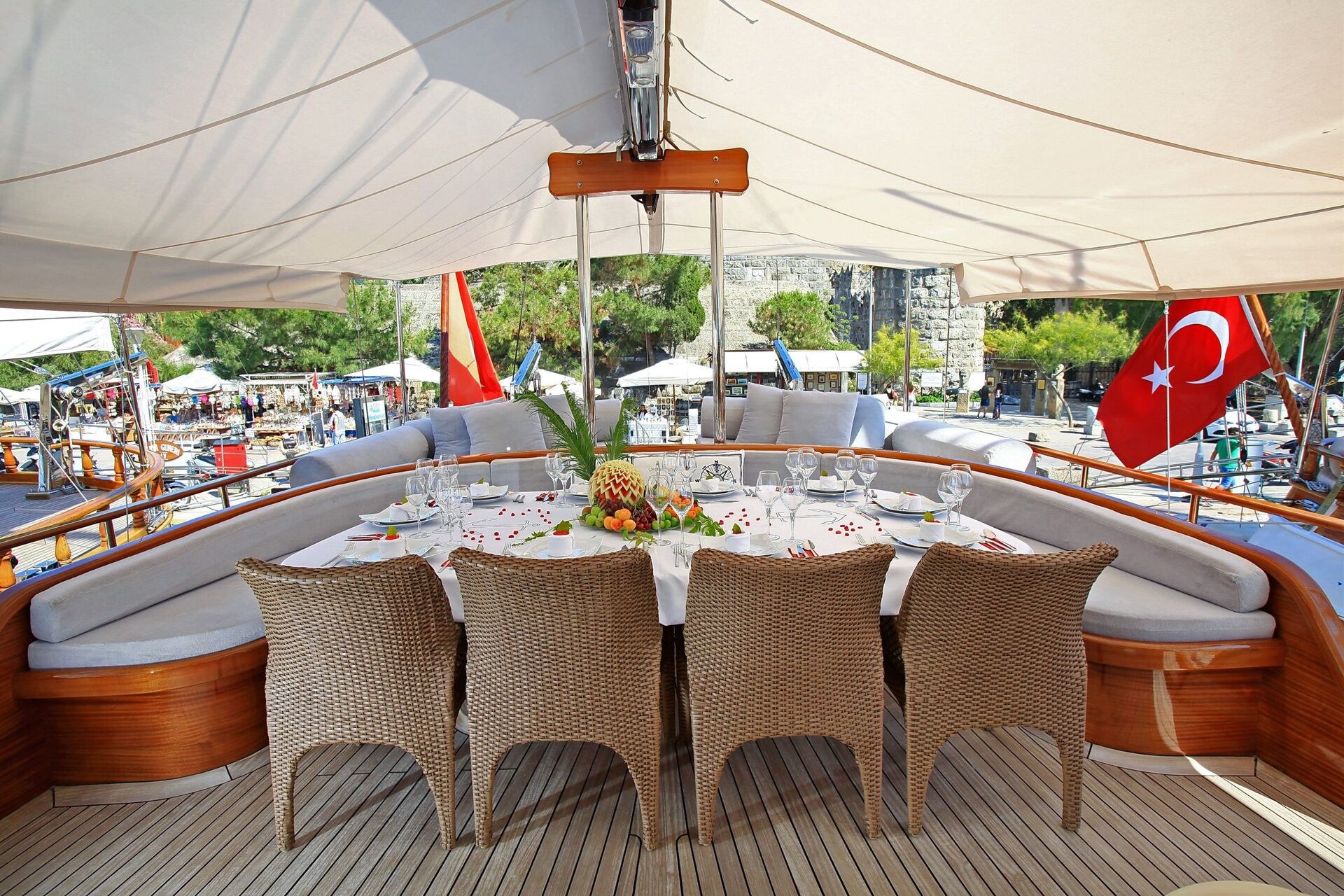 A dining table set for six with fruit centerpiece on a yacht deck, shaded by a canopy, with the Turkish flag and harbor in the background.