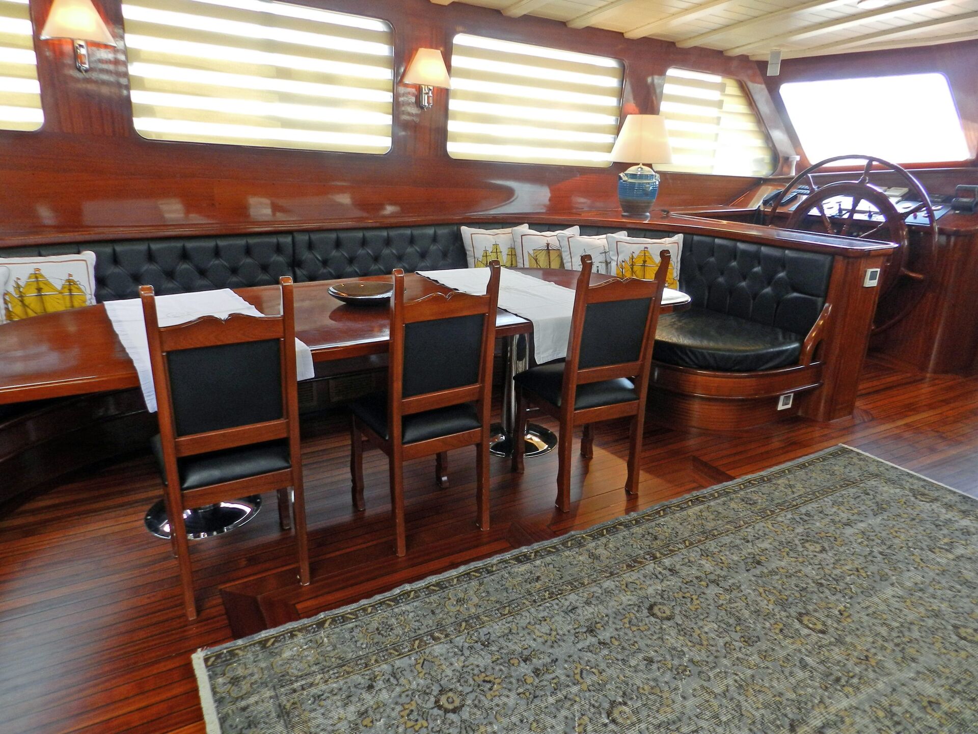 Elegant yacht interior with polished wooden floors, a dining table with four chairs, cushioned bench seating, nautical-themed pillows, lamps, and a steering wheel by large windows with blinds. Rug covers part of the floor.