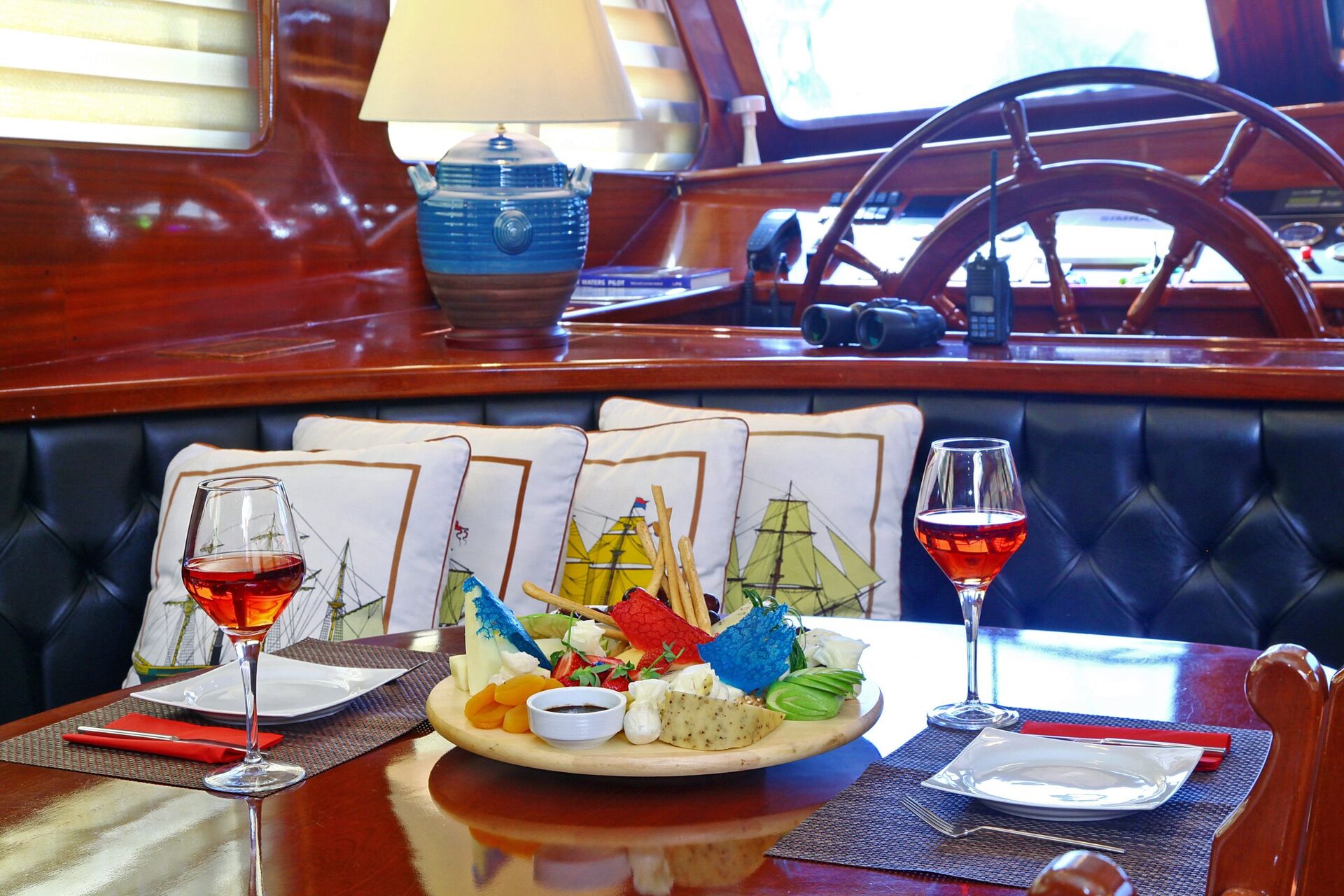 A table on a yacht set with two glasses of rosé wine, plates, and a large platter of assorted cheeses and fruits. Behind the table are nautical-themed pillows, a blue lamp, and a wooden steering wheel.