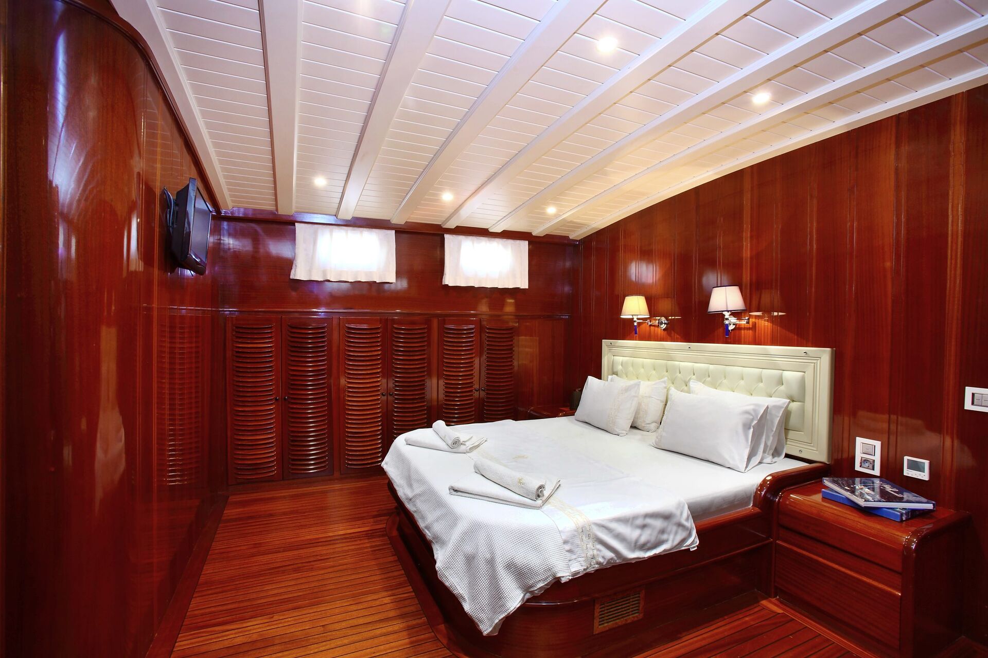 A cozy bedroom with polished wooden walls and floors, a double bed with white linens, two bedside tables, wall lamps, a TV on the wall, and small windows letting in natural light.