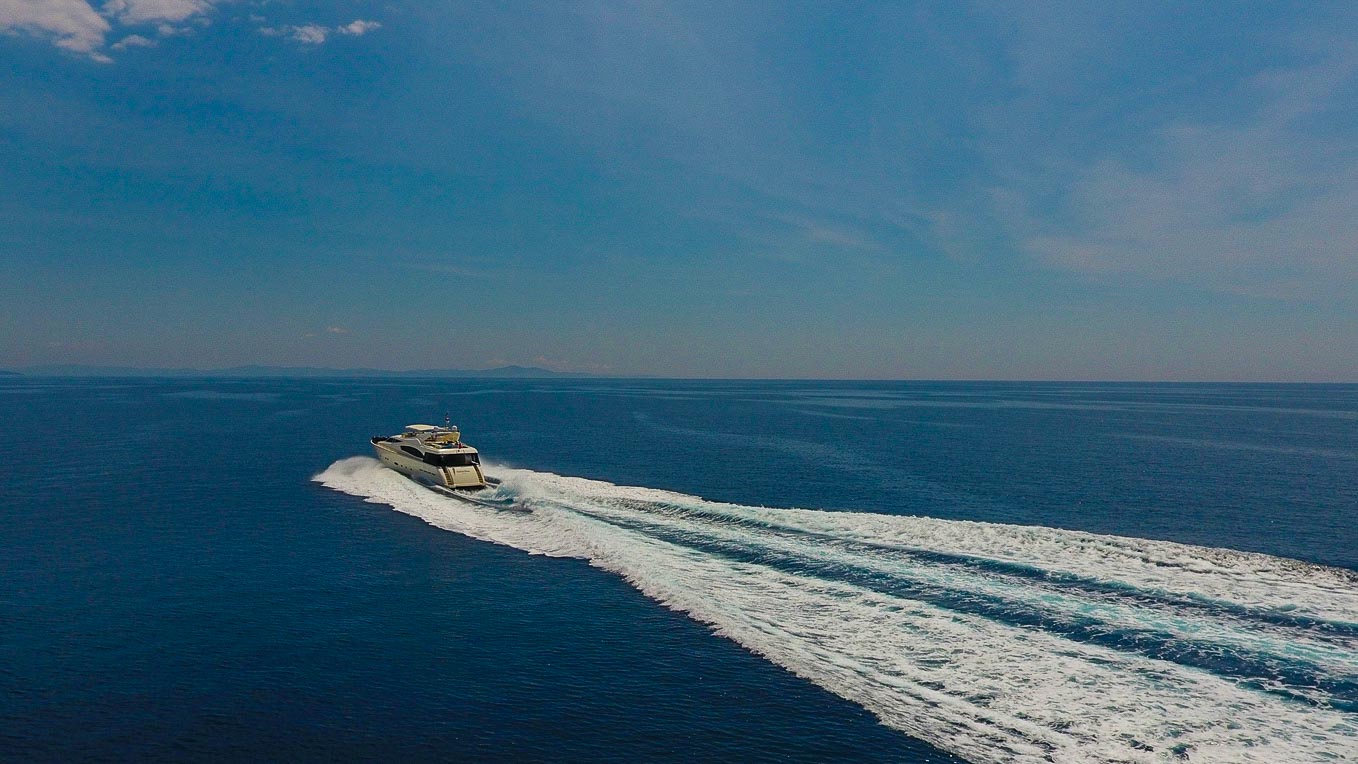 A yacht speeds across a calm blue sea, leaving a wide white wake behind it under a clear, sunny sky.
