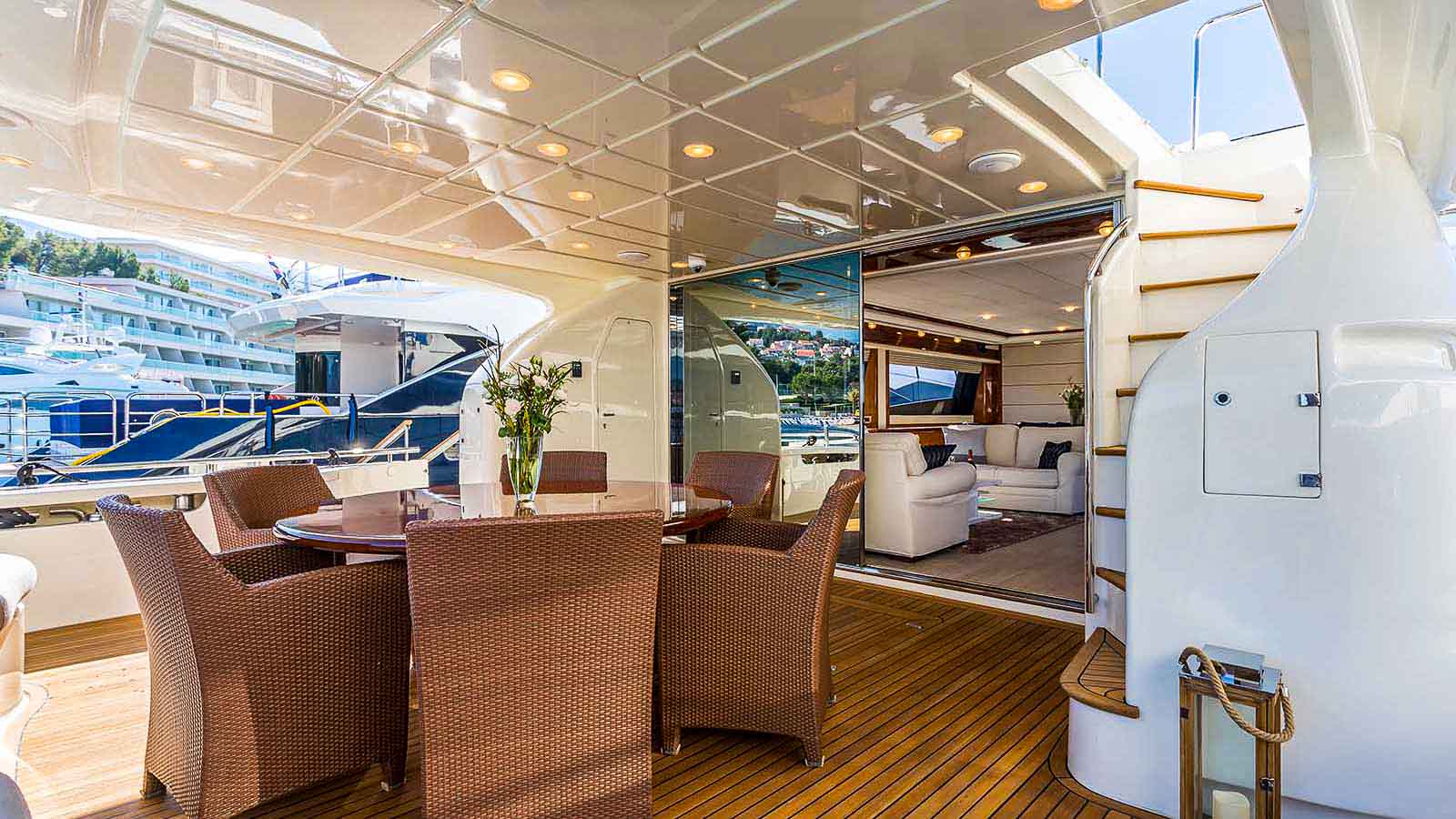 A luxurious yacht deck features a round dining table with six wicker chairs, a vase of flowers, wooden flooring, and stairs leading up. Sliding glass doors open to a modern lounge area inside the yacht.