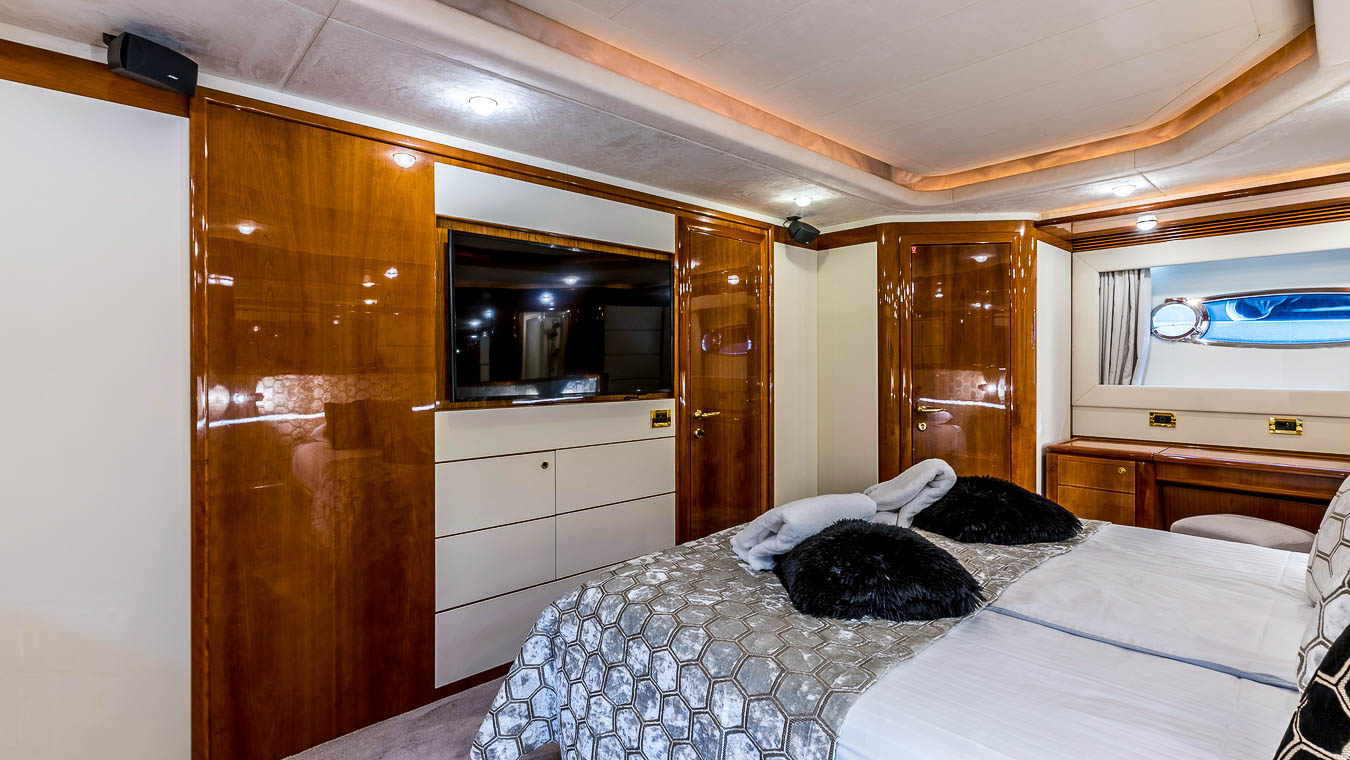 A luxurious yacht bedroom with glossy wood paneling, a wall-mounted TV, white drawers, a neatly made bed with patterned bedding and black pillows, and a window showing the outside.