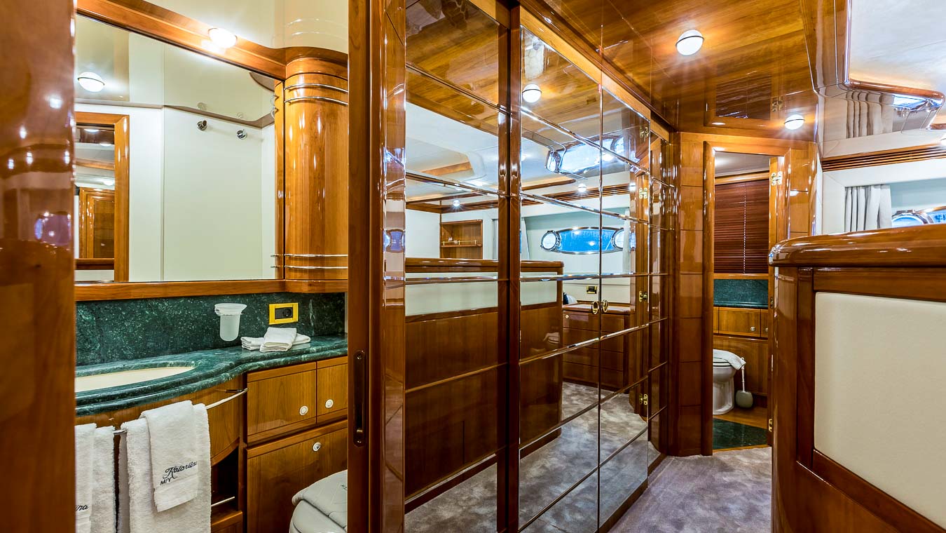 Luxurious yacht bathroom with glossy wood paneling, green marble countertops, mirrors, towels, and modern lighting. A sink and toilet are visible, creating an elegant and spacious atmosphere.
