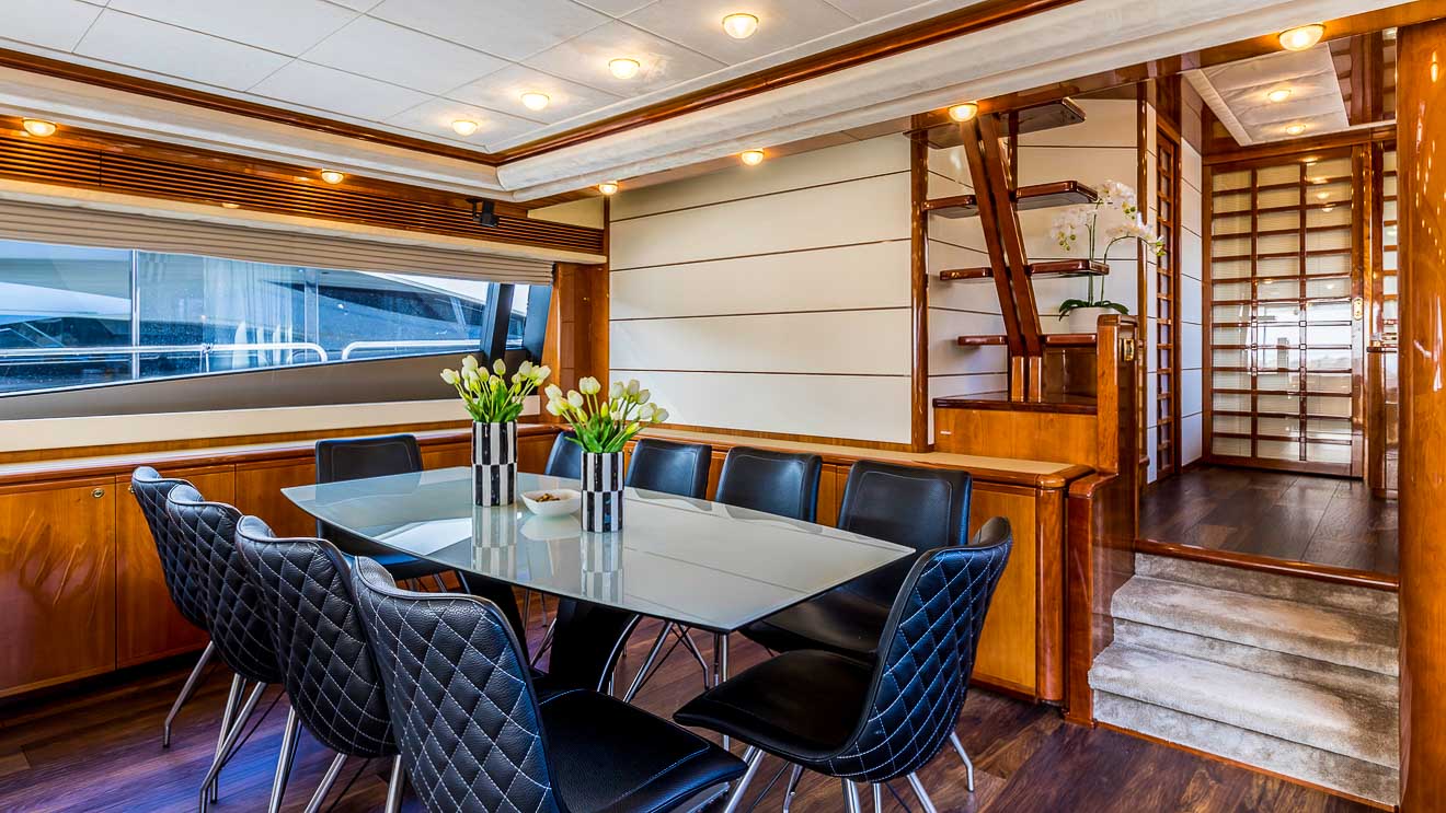 Modern dining area on a yacht with a glass table, eight black quilted chairs, wooden finishes, vases of white tulips, and large windows; stairs lead up to another level.
