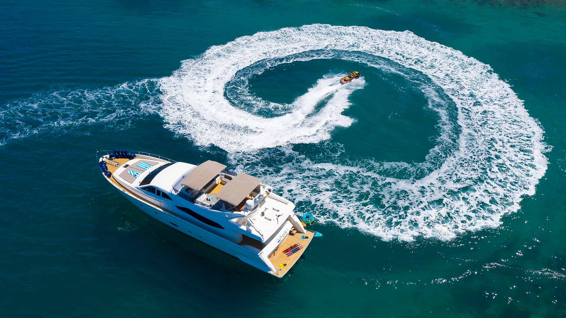 Aerial view of a large white yacht on blue water with a person riding a jet ski, creating a wide circular wake around the yacht.