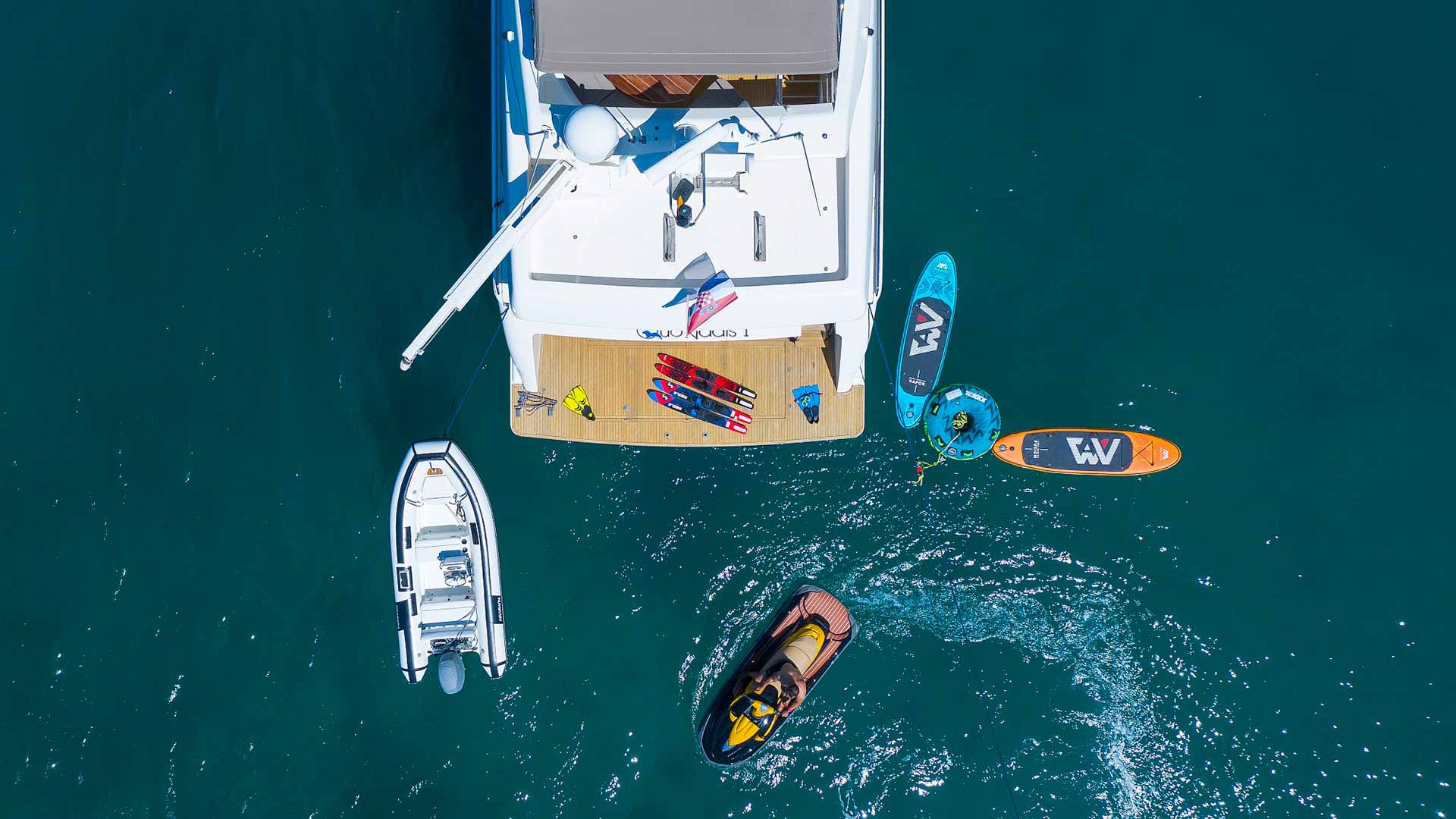 Aerial view of a yacht with water sports equipment on the deck, a small boat, a jet ski, and two paddleboards floating nearby in clear blue water.