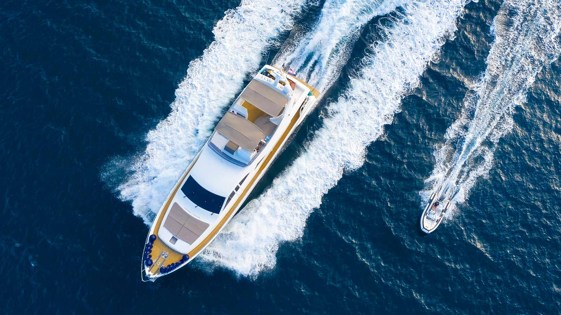 Aerial view of a large yacht cruising through deep blue water, creating white waves, with a smaller speedboat trailing closely behind on the right.