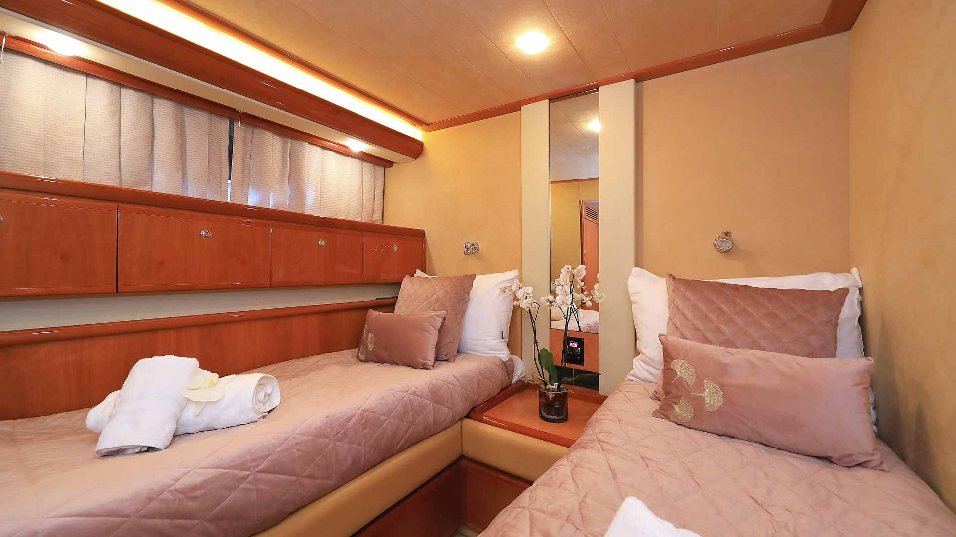 A cozy yacht cabin features two single beds with beige bedding, pillows, and a rolled towel on one bed. A small table with flowers is between the beds, and wood cabinets line the wall. Soft lighting creates a warm atmosphere.