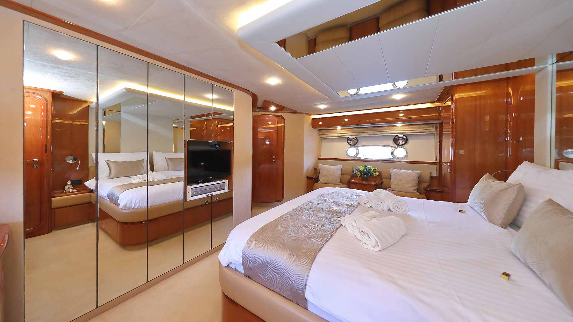 Luxurious yacht bedroom with wooden accents, mirrored closet doors, a large bed with neatly arranged towels, beige bedding, and windows offering natural light. A TV and seating area are also visible.