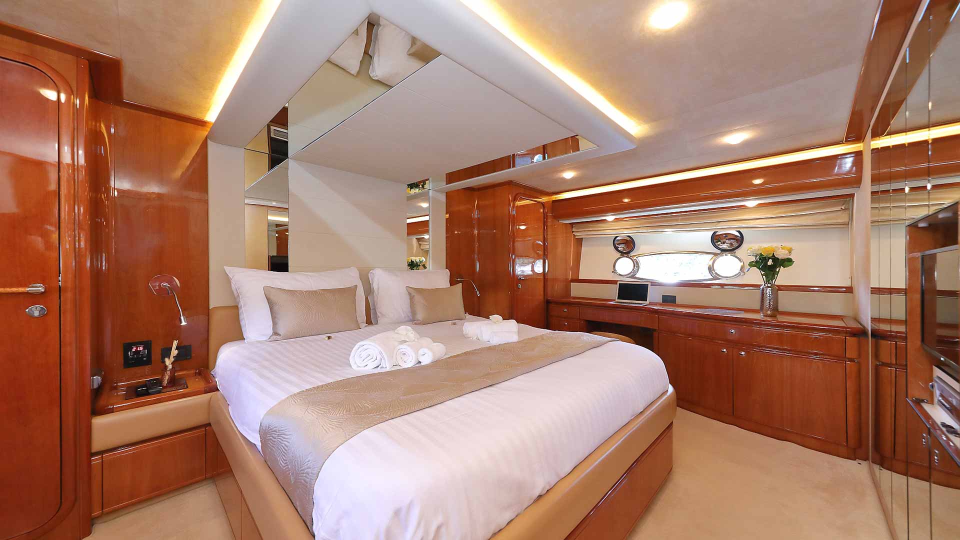A luxurious yacht bedroom features a large bed with crisp white linens, beige accents, rolled towels, wooden cabinets, fresh flowers, and windows with circular portholes letting in natural light.