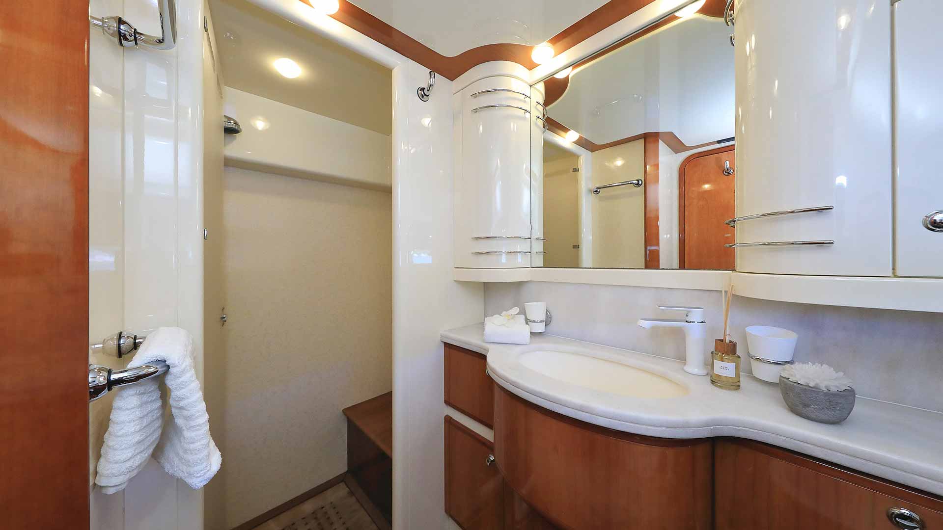Modern yacht bathroom with glossy wood and white cabinets, a curved countertop with sink, a towel on a hook, toiletries, and a walk-in shower in the background under recessed lighting.