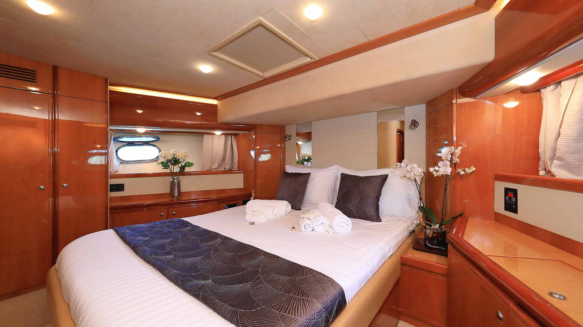 A well-lit yacht cabin features a neatly made double bed with dark and white pillows, rolled towels, wood-paneled walls and cabinets, a vase of flowers, and small windows with curtains.
