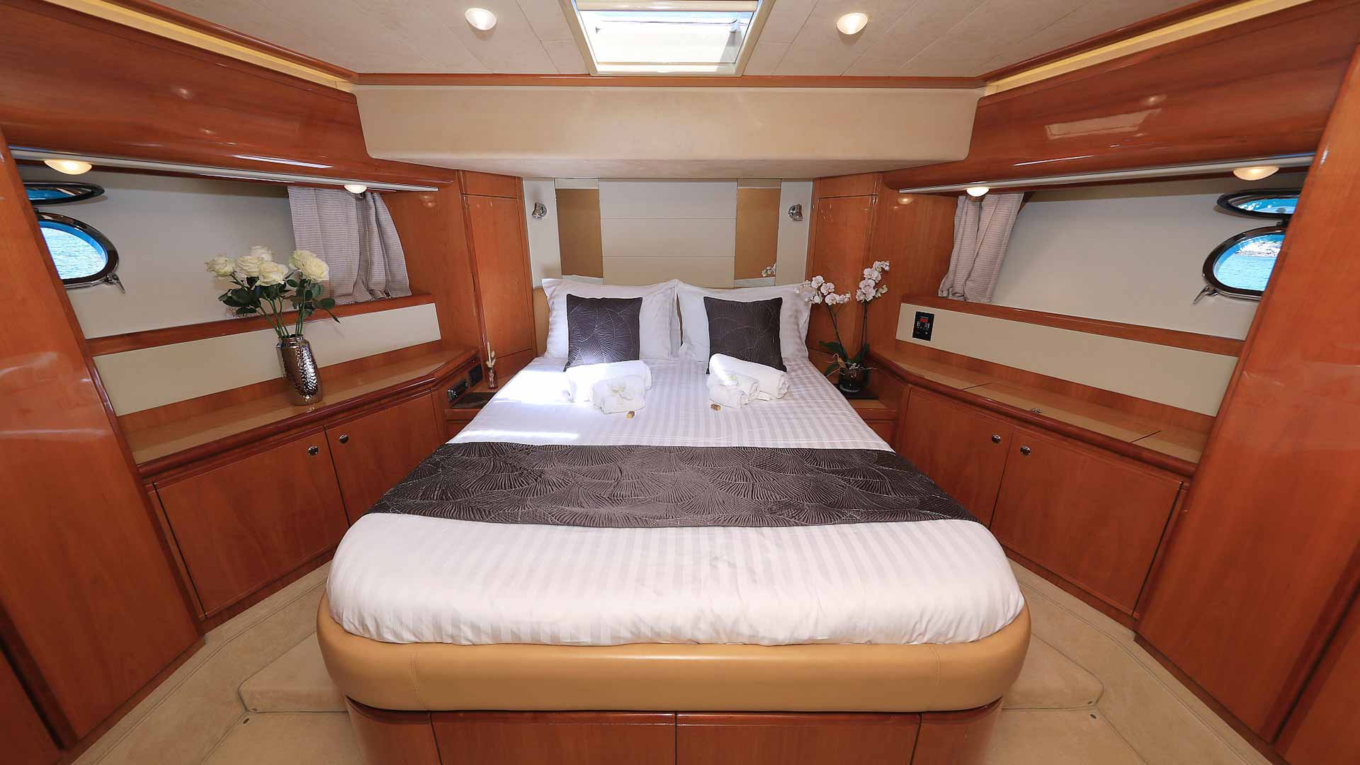 A well-lit yacht bedroom features a neatly made bed with white linens, gray accents, and decorative pillows, surrounded by wooden cabinetry, vases with flowers, and small porthole windows on either side.
