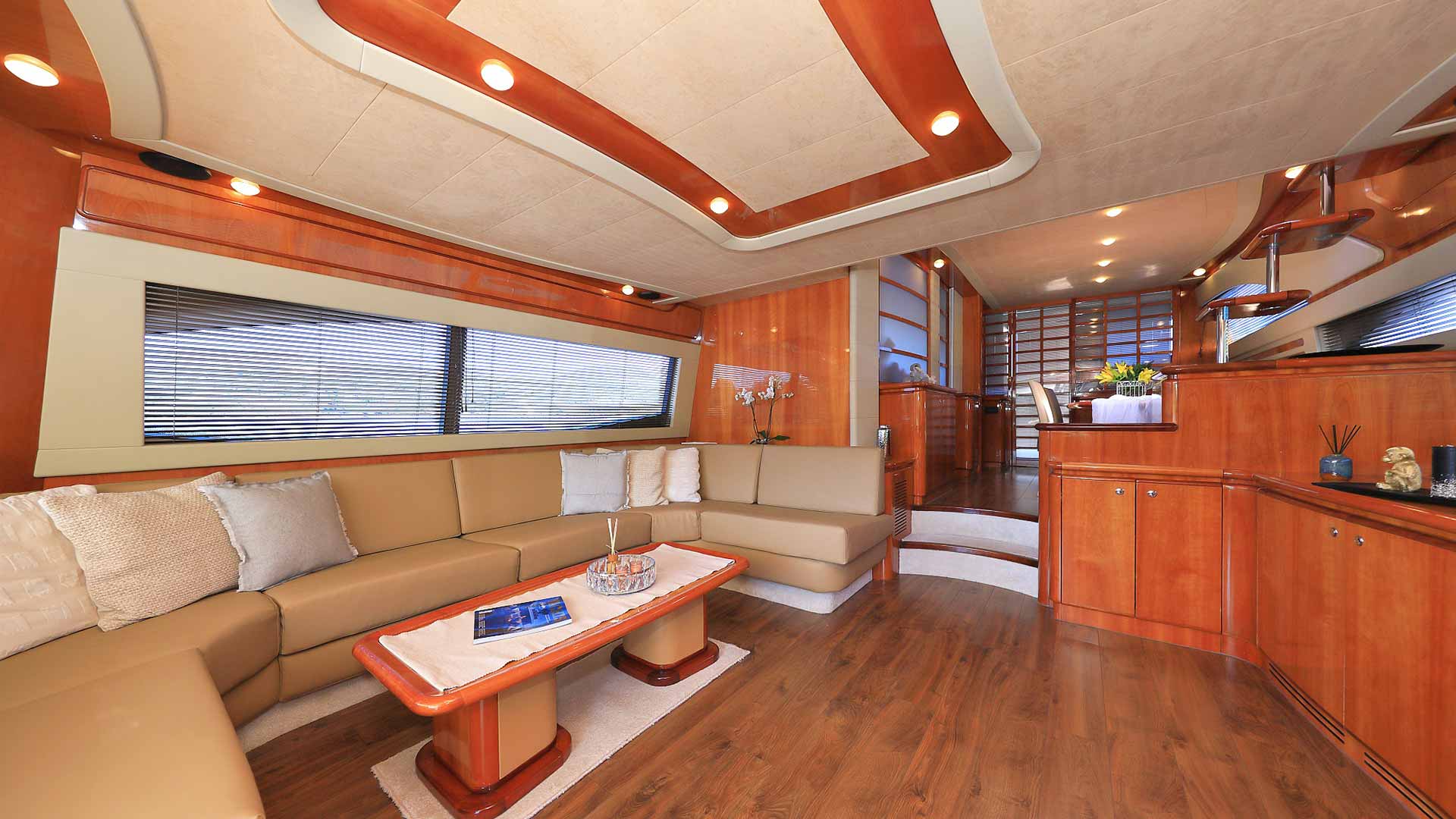 Luxurious yacht interior with cream-colored L-shaped sofa, wood floors, glossy wood walls, rectangular coffee table, decorative pillows, and large windows with blinds, leading to a dining area in the background.