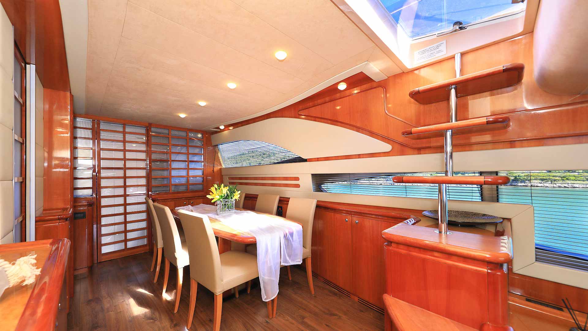 A modern yacht interior with wooden paneling, a dining table set for six with a vase of yellow flowers, cream chairs, large windows, and a spiral staircase leading to an upper deck. Sunlight streams in through a skylight.