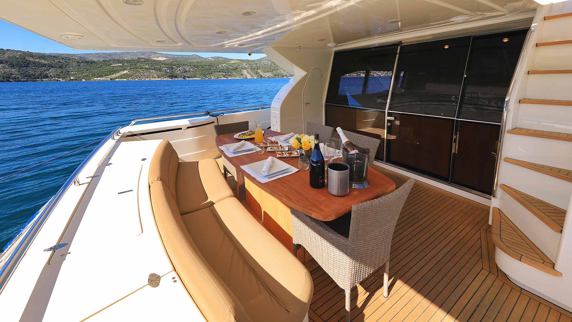 A luxurious yacht deck features a dining table set for four with wine, fruit, and breakfast on plates. Cushioned seating and wicker chairs face clear blue water and distant green hills under a sunny sky.