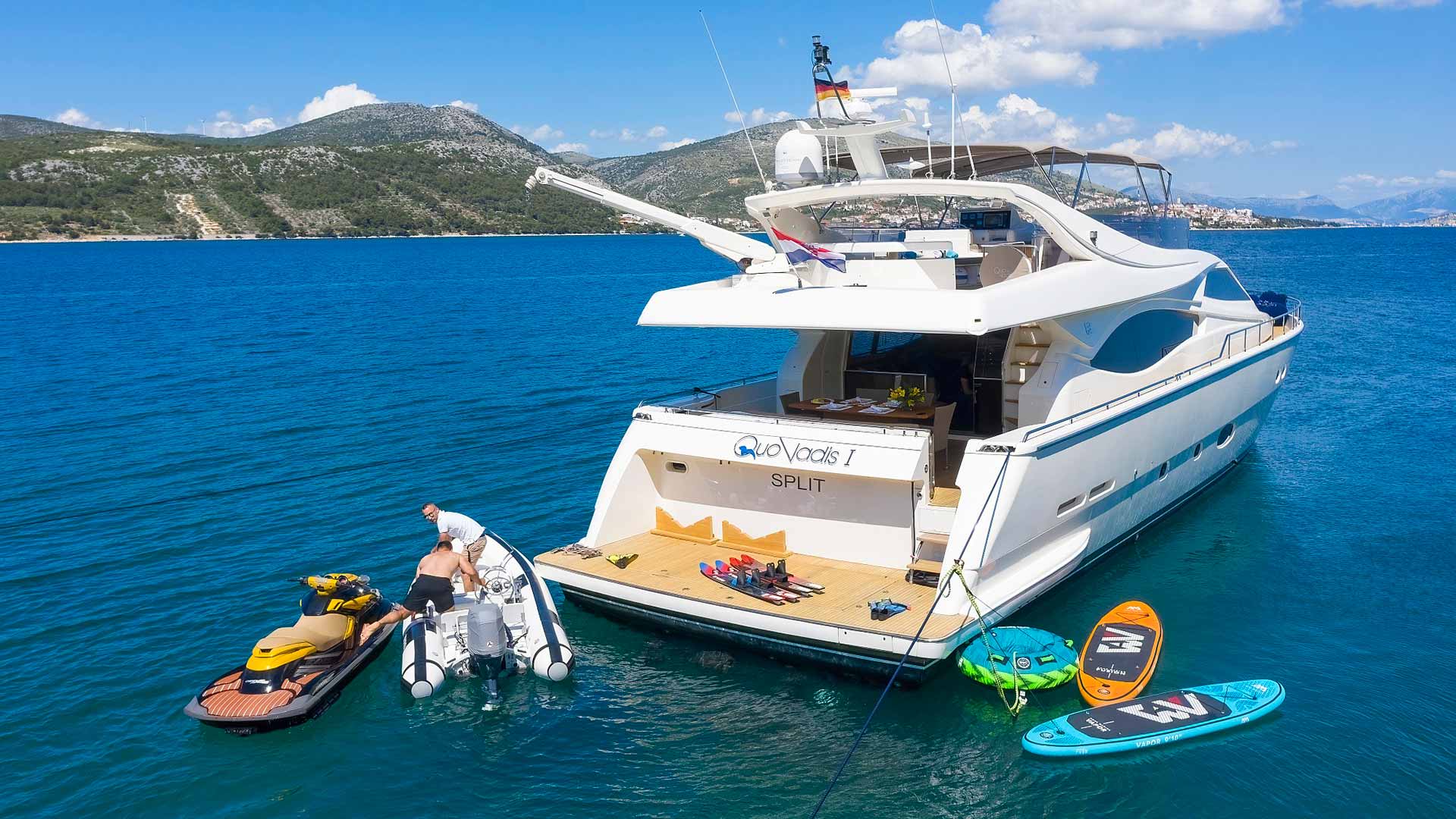 A white yacht named Split is anchored in clear blue water near a mountainous coastline. A person stands on a small inflatable boat beside a jet ski, with paddleboards and water toys on the yacht’s rear deck.