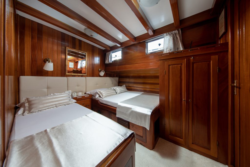 A cozy cabin room with wood-paneled walls and ceiling, featuring two beds with white linens, a wardrobe, a mirror, small windows, and wall-mounted lamps, creating a warm and inviting atmosphere.