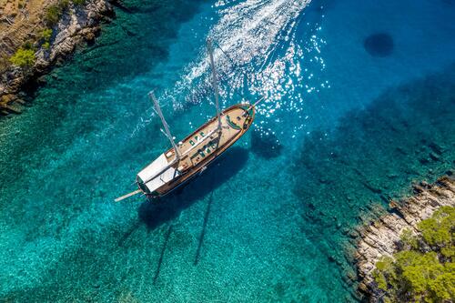 Aerial view of a wooden sailboat gliding through clear blue water near rocky, tree-lined shores, with sunlight sparkling on the seas surface.