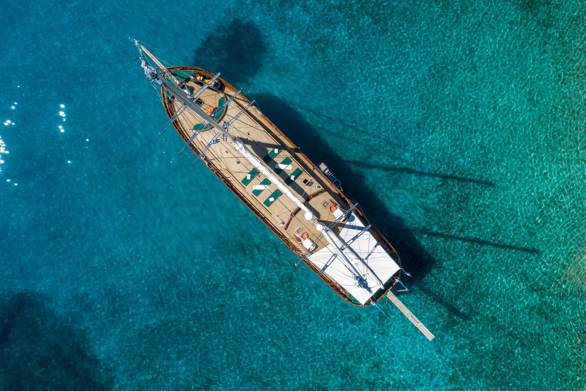 Aerial view of a wooden sailboat with sun loungers on its deck, floating on clear, turquoise water. The boat casts a shadow in the shallow, transparent sea below.