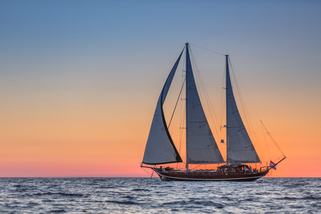 Un velero con dos mástiles altos se desliza por aguas tranquilas al atardecer, silueteado contra un cielo vibrante de tonos naranjas, rosas y azules.
