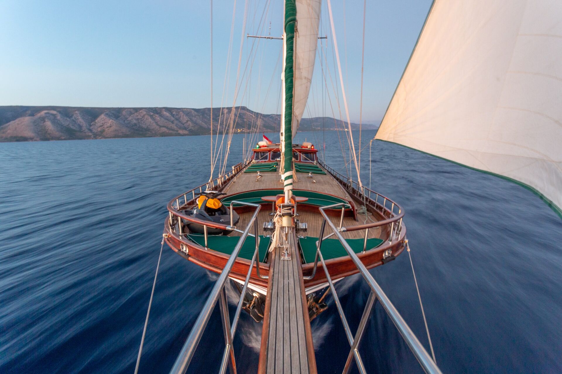 A view from the bow of a sailboat sailing on calm blue water, with sails unfurled and distant mountains visible on the horizon under a clear sky.